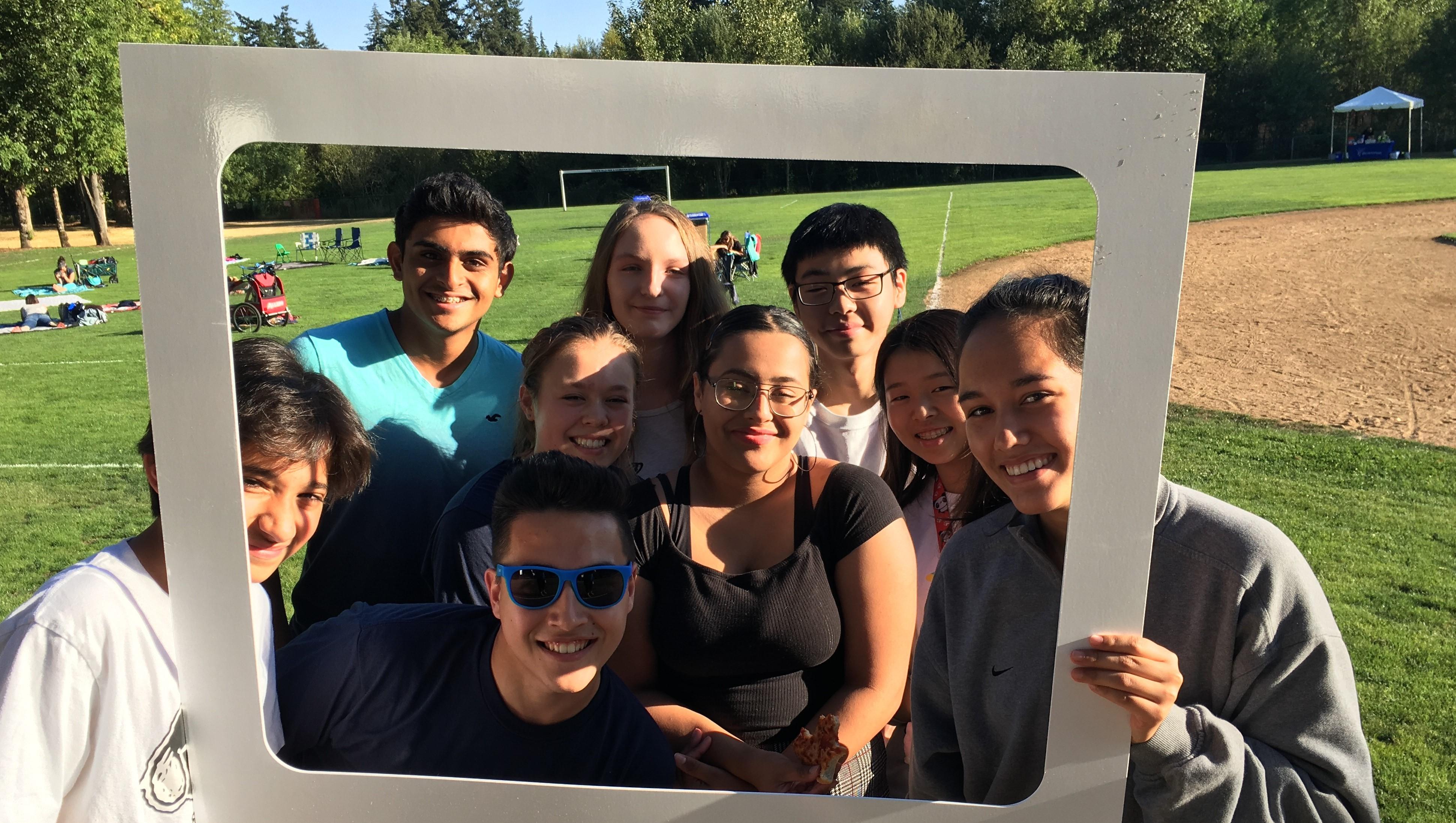 A group of nine individuals posing cheerfully behind a large, handheld gray frame in a sunny park setting.