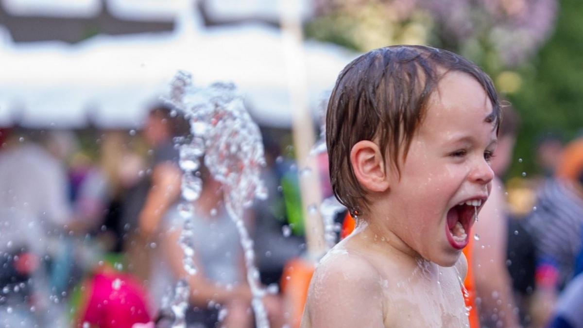 A young child laughing with joy as water splashes around him at an outdoor event, with blurred figures of other people in the background.